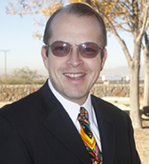 photo of Tom Laign wearing a dark suit with a a multi-colored tie, white shirt, poloarized eye glasses standing before a yard